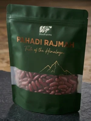 A close-up photograph of a premium, matte forest-green stand-up pouch containing HimFarms Pahadi Rajmah. The packaging features elegant rose gold foil typography reading "Pahadi Rajmah - Taste of the Himalayas," a gold line-art mountain illustration, and a clear transparent window across the lower half showing the raw red kidney beans inside. The pouch rests on a dark grey slate stone surface against a blurred rustic wooden background.