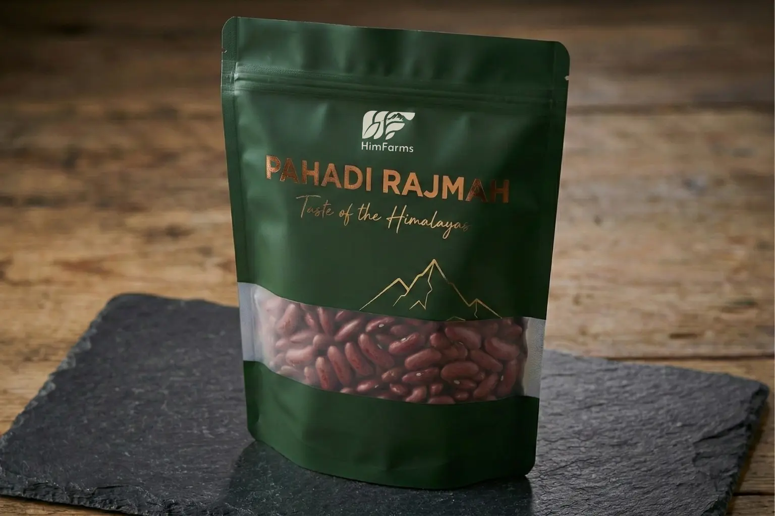 A close-up photograph of a premium, matte forest-green stand-up pouch containing HimFarms Pahadi Rajmah. The packaging features elegant rose gold foil typography reading "Pahadi Rajmah - Taste of the Himalayas," a gold line-art mountain illustration, and a clear transparent window across the lower half showing the raw red kidney beans inside. The pouch rests on a dark grey slate stone surface against a blurred rustic wooden background.